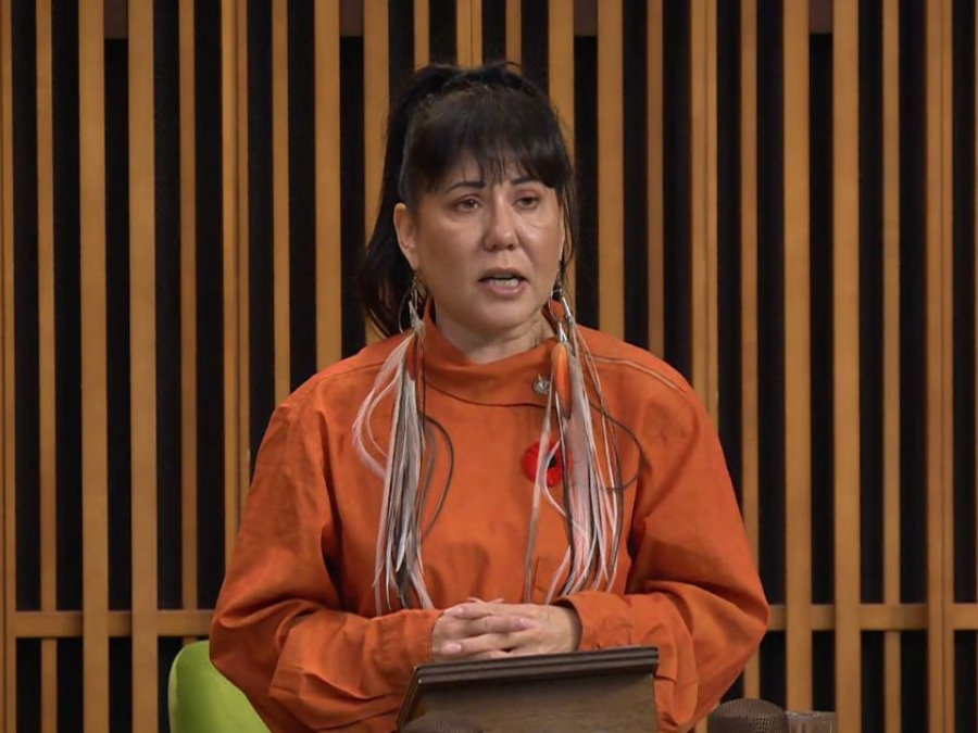 A woman stands at a podium, wearing a bright orange top and feather earrings, speaking earnestly. A poppy is pinned to her chest, symbolizing remembrance.