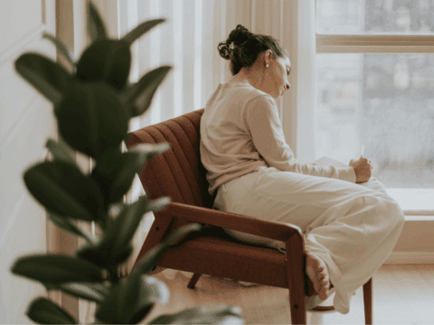 A woman in a light sweater and white pants sits curled up on a chair by a window, thoughtfully writing. A large green plant is in the foreground.