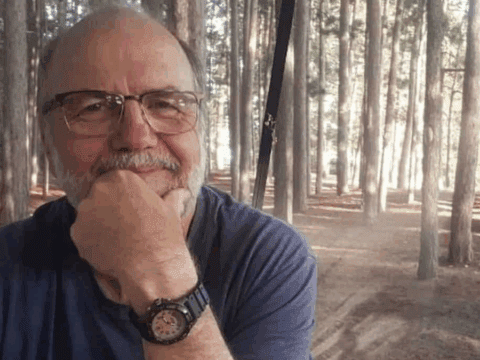A man with glasses and a watch smiles gently with his hand on his chin, seated in a serene pine forest. Sunlight filters through the trees, creating a calm atmosphere.