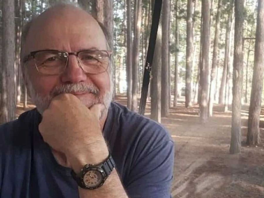A man with glasses and a watch smiles gently with his hand on his chin, seated in a serene pine forest. Sunlight filters through the trees, creating a calm atmosphere.