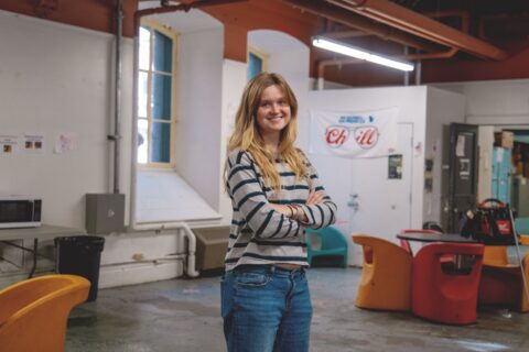 Smiling woman with long hair, in a striped sweater and jeans, stands confidently with arms crossed in a casual room featuring colorful chairs and a "Chill" banner.