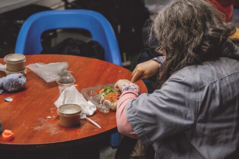 A person with long hair is seated at a round table with a blue chair, eating from a plastic container. The table holds two soup cups and napkins. The atmosphere is casual.