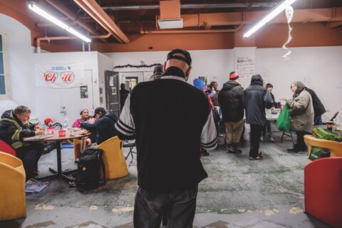 Community shelter interior with people gathered. Some sit and eat at tables, others stand in line. Warm lighting creates a welcoming atmosphere.