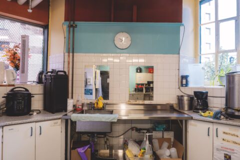 A tidy kitchen with a sink and countertop featuring cleaning supplies, a coffee maker, and a slow cooker. A wall clock hangs above. Sunlight enters through a window.