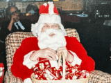 Santa, in bright red and white suit, sits on a patterned chair holding a basket with red and green wrapped gifts. A man sits in the background. Cozy holiday scene.