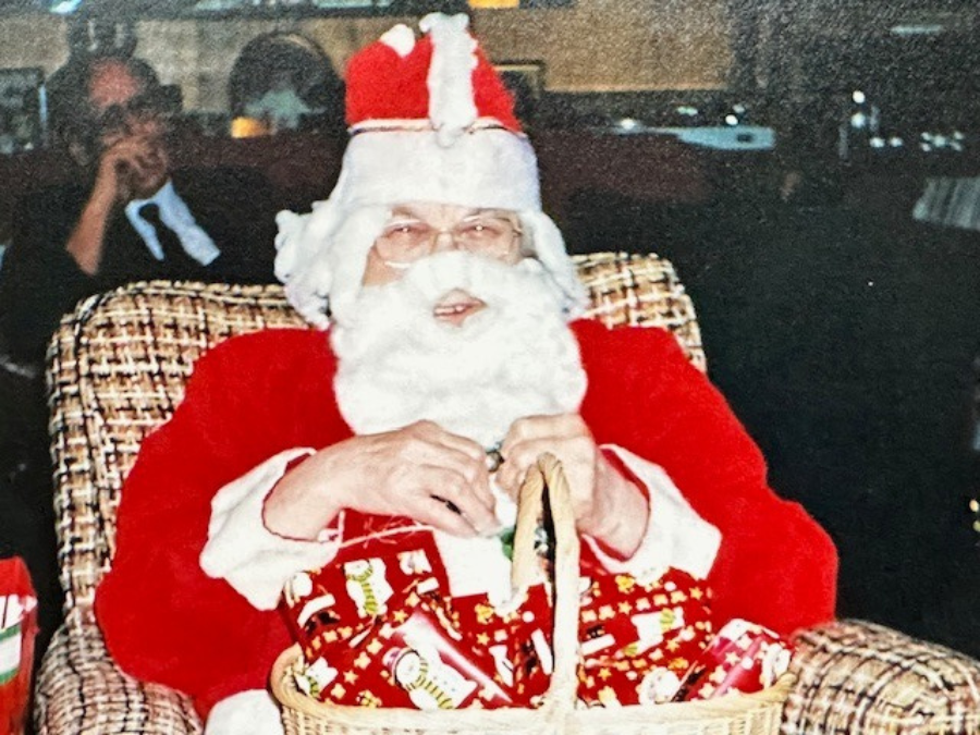 Santa, in bright red and white suit, sits on a patterned chair holding a basket with red and green wrapped gifts. A man sits in the background. Cozy holiday scene.