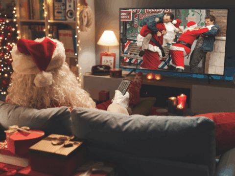 Santa in a cozy room watches a Christmas movie, featuring festive chaos, on TV. Surroundings include a lit tree, gifts, and warm candlelight.