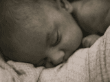 A sepia-toned image of a newborn baby peacefully sleeping on a soft, textured blanket. The atmosphere is serene and tender, evoking warmth and comfort.