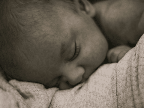 A sepia-toned image of a newborn baby peacefully sleeping on a soft, textured blanket. The atmosphere is serene and tender, evoking warmth and comfort.