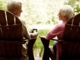 Elderly couple sitting in wooden chairs on a porch, facing each other. They overlook a lush, green landscape, conveying a peaceful and serene mood.