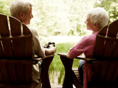 Elderly couple sitting in wooden chairs on a porch, facing each other. They overlook a lush, green landscape, conveying a peaceful and serene mood.