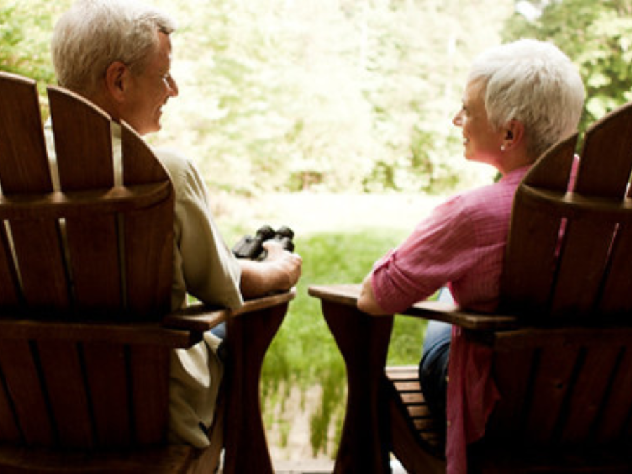 Elderly couple sitting in wooden chairs on a porch, facing each other. They overlook a lush, green landscape, conveying a peaceful and serene mood.