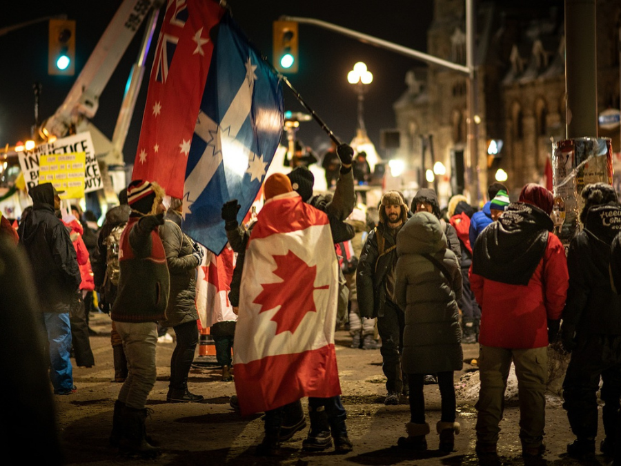 Crowd of protesters at night, waving Canadian and other flags. Participants wear winter clothing, creating a lively, determined atmosphere with glowing streetlights.