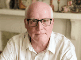A man with glasses and a white shirt sits indoors, looking directly at the camera with a calm expression. Background shows blurred decor on a shelf.