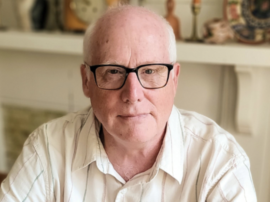 A man with glasses and a white shirt sits indoors, looking directly at the camera with a calm expression. Background shows blurred decor on a shelf.