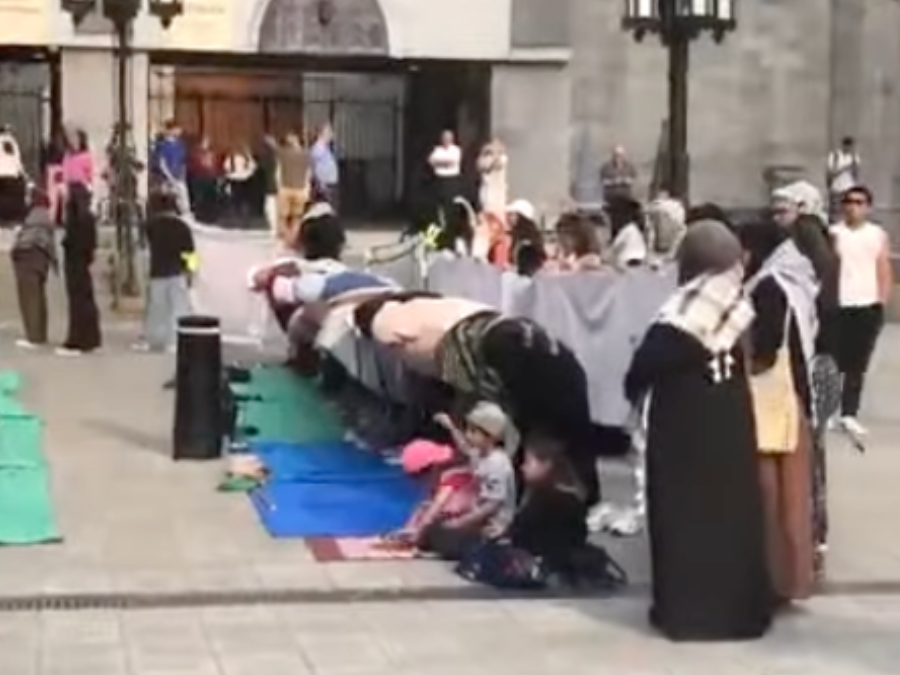 Outdoor gathering with people in prayer, some kneeling on colorful mats. Others stand nearby, watching. The mood is peaceful and reverent.
