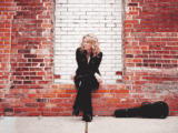 Woman in black outfit sits contemplatively on a rustic red-brick wall with a white section. Guitar case lies beside her, contrasting urban vibe.