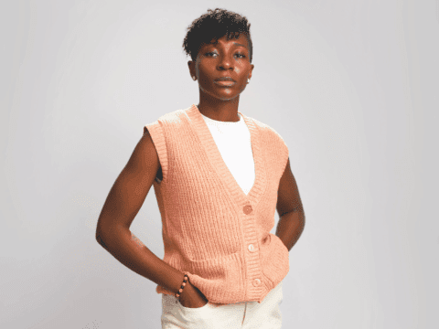 A person in a peach knitted vest and white shirt with hands in pockets. They stand confidently against a plain background, conveying a calm, poised demeanor.