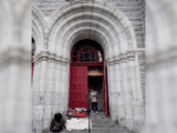 A person enters a large stone building through an open red door. Blankets and bags are stacked nearby, suggesting homeless shelter or aid context.