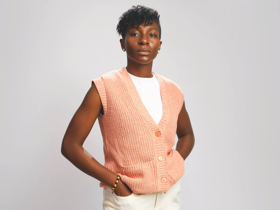 A person in a peach knitted vest and white shirt with hands in pockets. They stand confidently against a plain background, conveying a calm, poised demeanor.