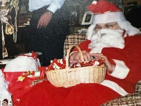 Santa, in bright red and white suit, sits on a patterned chair holding a basket with red and green wrapped gifts. A man sits in the background. Cozy holiday scene.