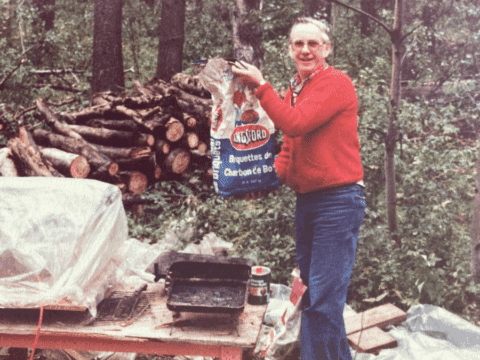 A person in a red sweater holds a charcoal bag next to a grill in a forest. Logs stacked behind them. The scene feels rustic and outdoorsy.