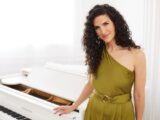 Musician Laila Biali stands in a green dress next to a white piano