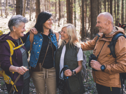 Four smiling adults stand in a forest, wearing hiking gear and backpacks. They embrace happily, conveying camaraderie and joy in nature.