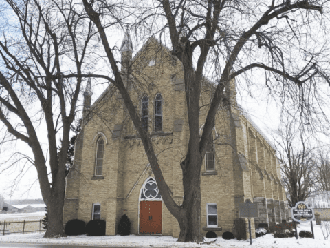 Historic stone church on a snowy day, featuring tall arched windows, a wooden door, and bare winter trees framing the entrance. Peaceful atmosphere.