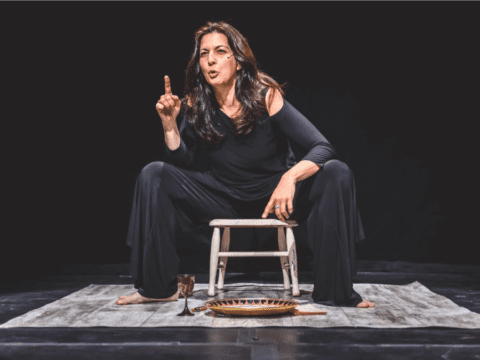 A woman in a flowing black outfit sits on a stool on stage, gesturing expressively. Her intense expression suggests a compelling performance.