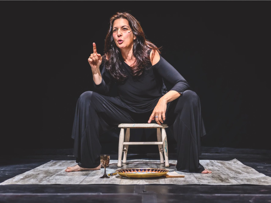 A woman in a flowing black outfit sits on a stool on stage, gesturing expressively. Her intense expression suggests a compelling performance.
