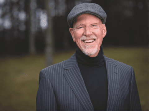 A man in a gray flat cap and striped suit jacket smiles warmly in an outdoor setting with blurred trees, conveying a friendly and relaxed tone.