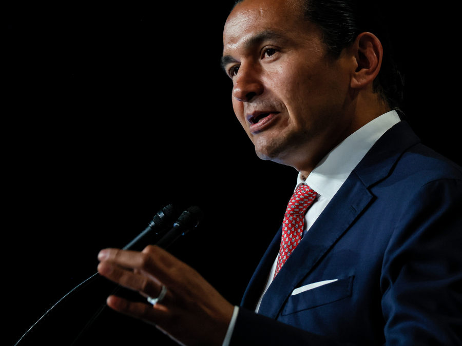 A man in a dark suit and red tie speaks emphatically at a microphone against a black background, conveying determination and focus.