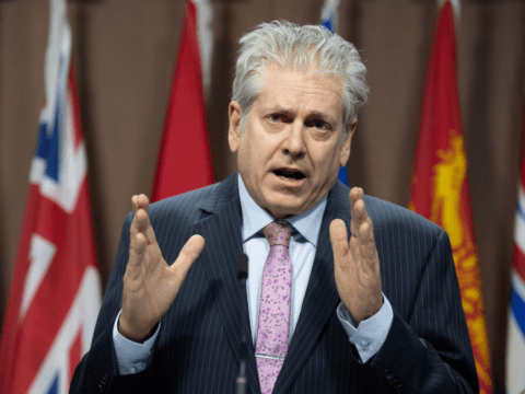 A man in a suit with a pink tie speaks passionately at a podium, gesturing with hands. Several flags are blurred in the background.