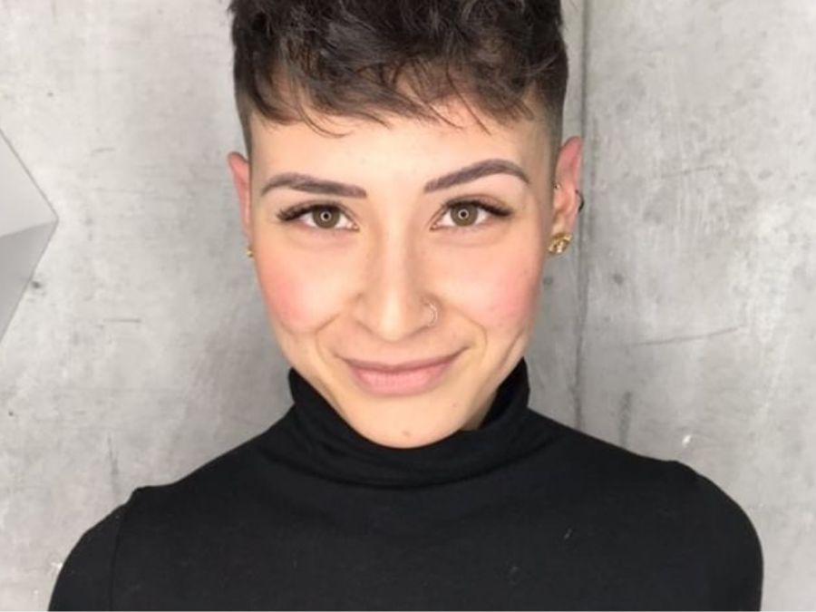 Smiling person with short, dark hair and a nose ring, wearing a black turtleneck, stands against a textured gray wall, exuding a confident and friendly demeanor.