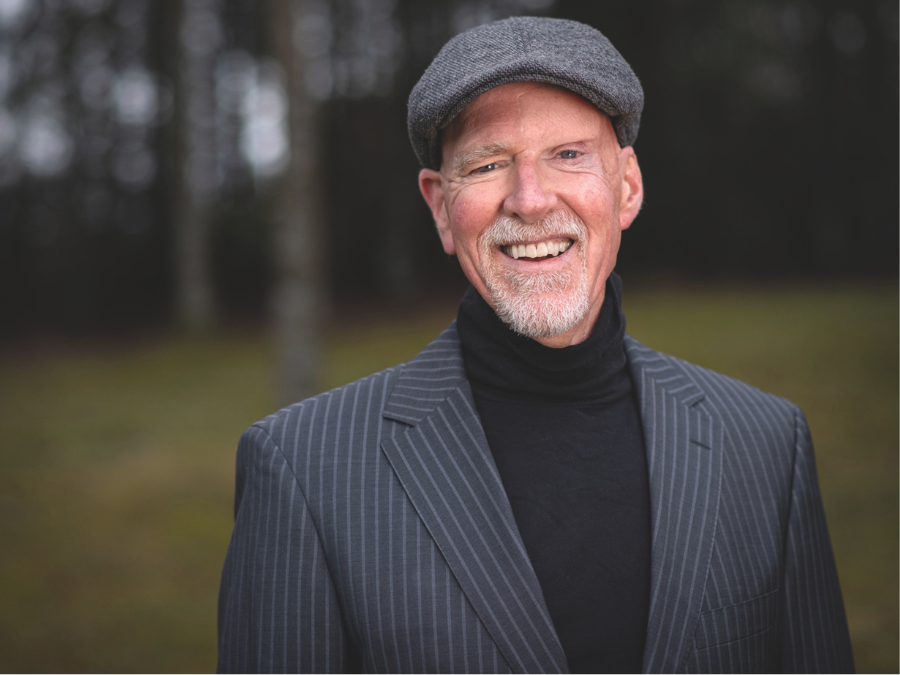 A man in a gray flat cap and striped suit jacket smiles warmly in an outdoor setting with blurred trees, conveying a friendly and relaxed tone.