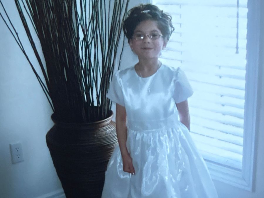 A girl with glasses and a white dress smiles for the camera in front of a plant
