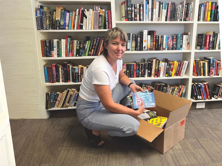 A Caucasian woman crouches on a floor next to a box of books. A bookcase stands behind her.