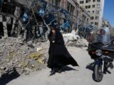 A woman walks past a damaged building. Next to her is a man on a motorcycle