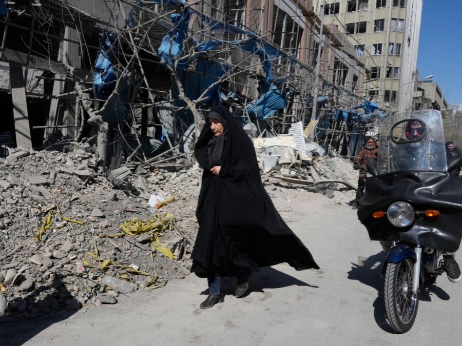 A woman walks past a damaged building. Next to her is a man on a motorcycle